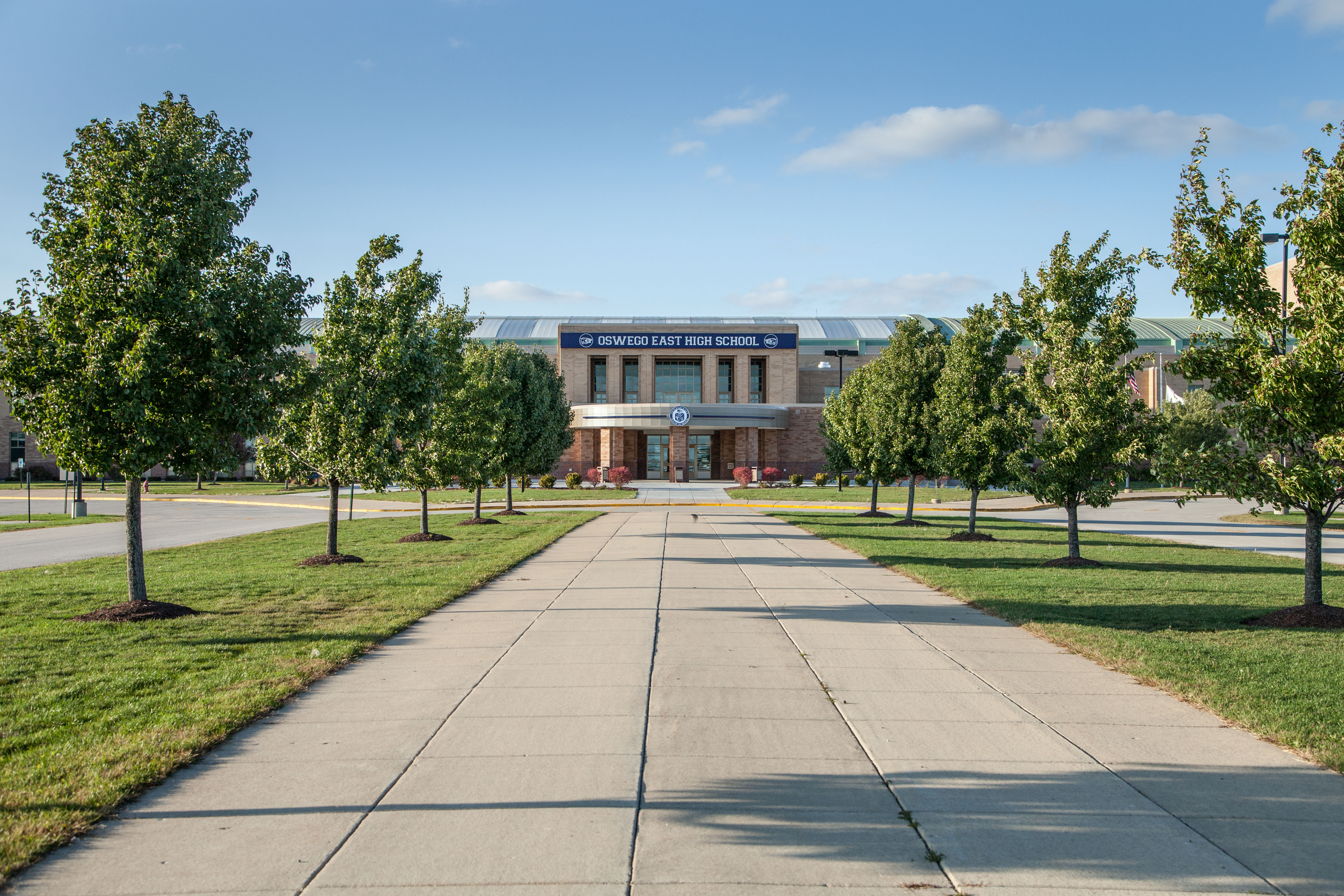 OEHS Entrance way highlighting a large center walkway with trees on both sides leading up to the building with a blue sky in the background.